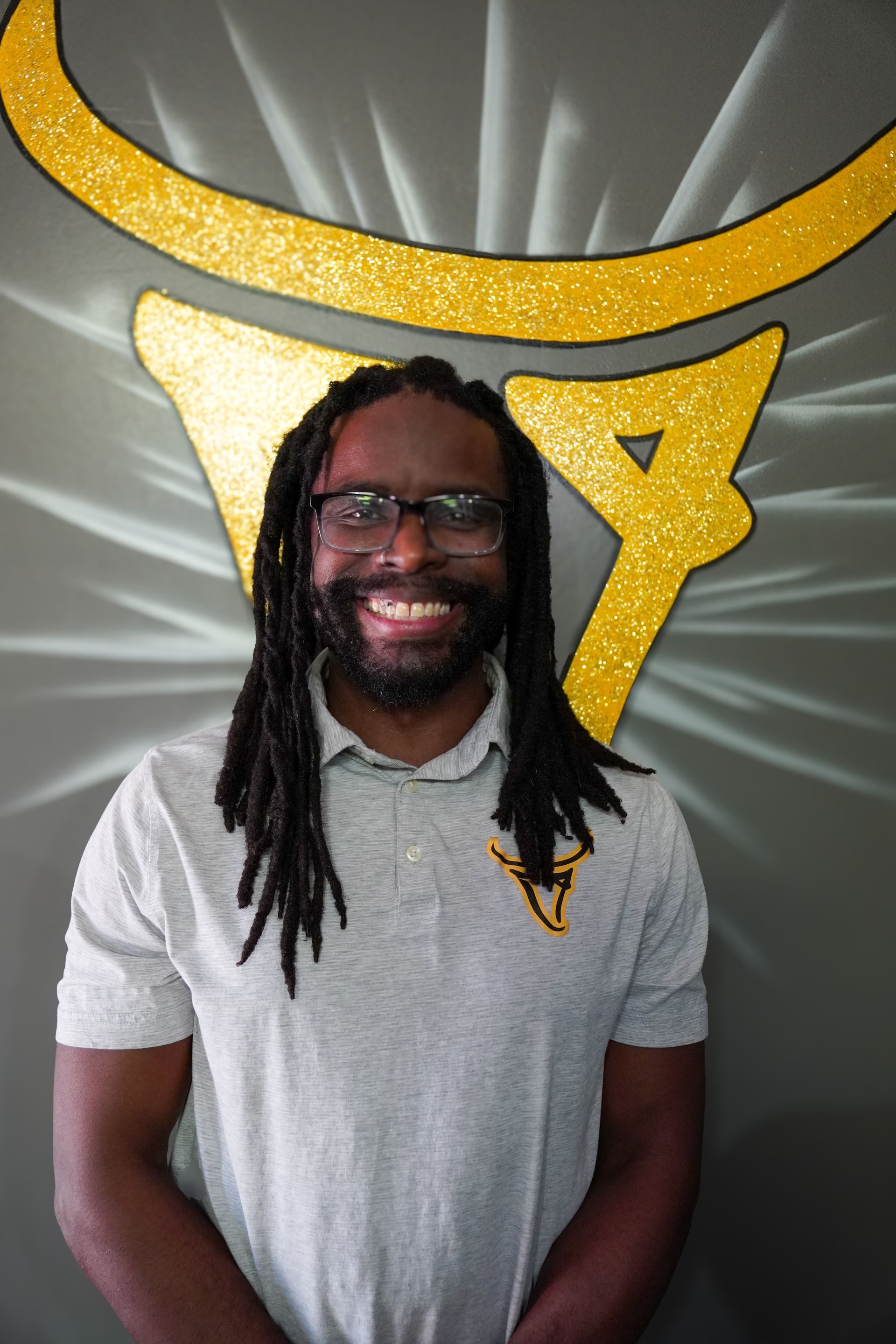 Man with dreadlocks and glasses smiles in front of a gold logo on a gray wall.