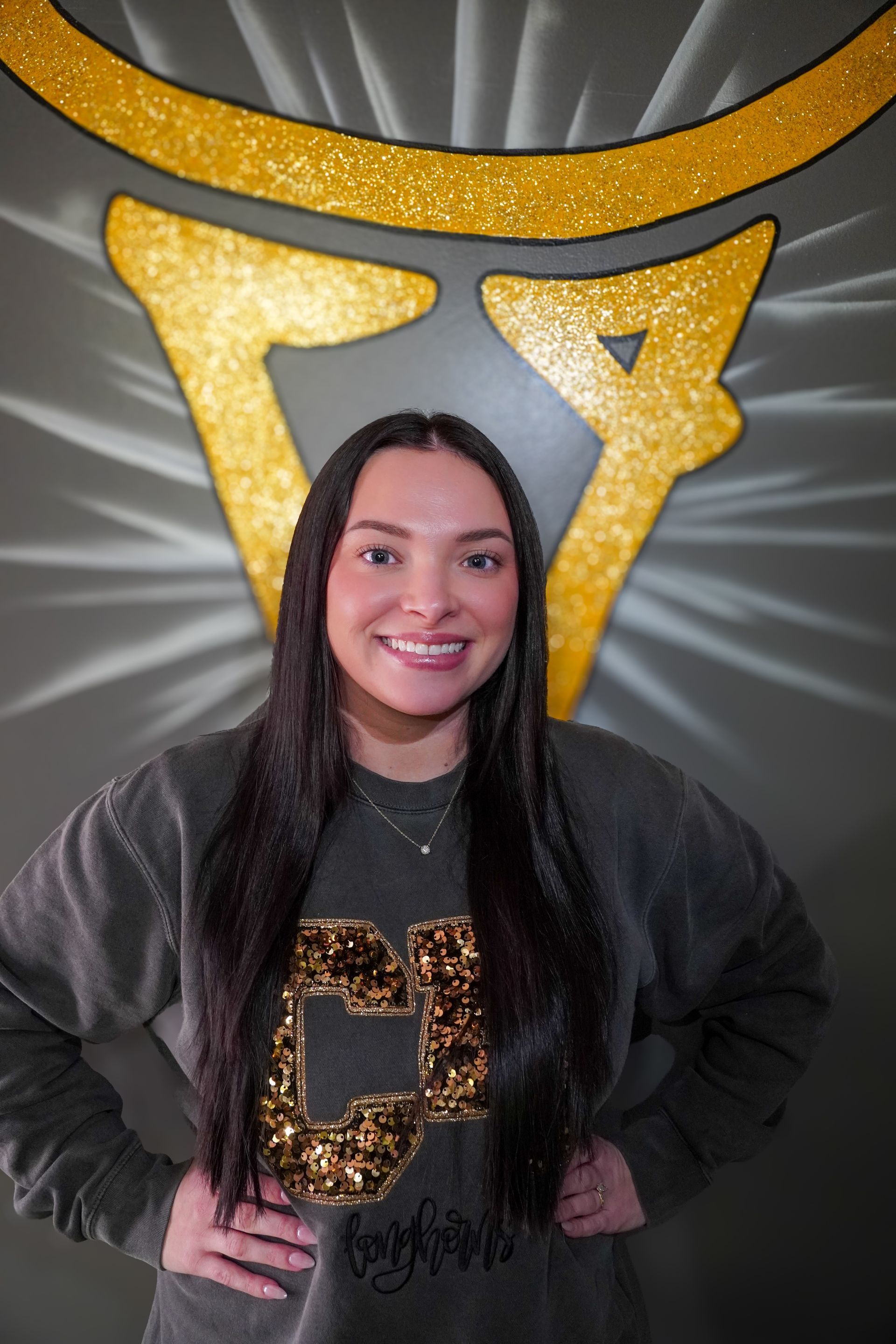 Woman with long dark hair, wearing a green sweatshirt, smiles in front of a gold, glittery logo.