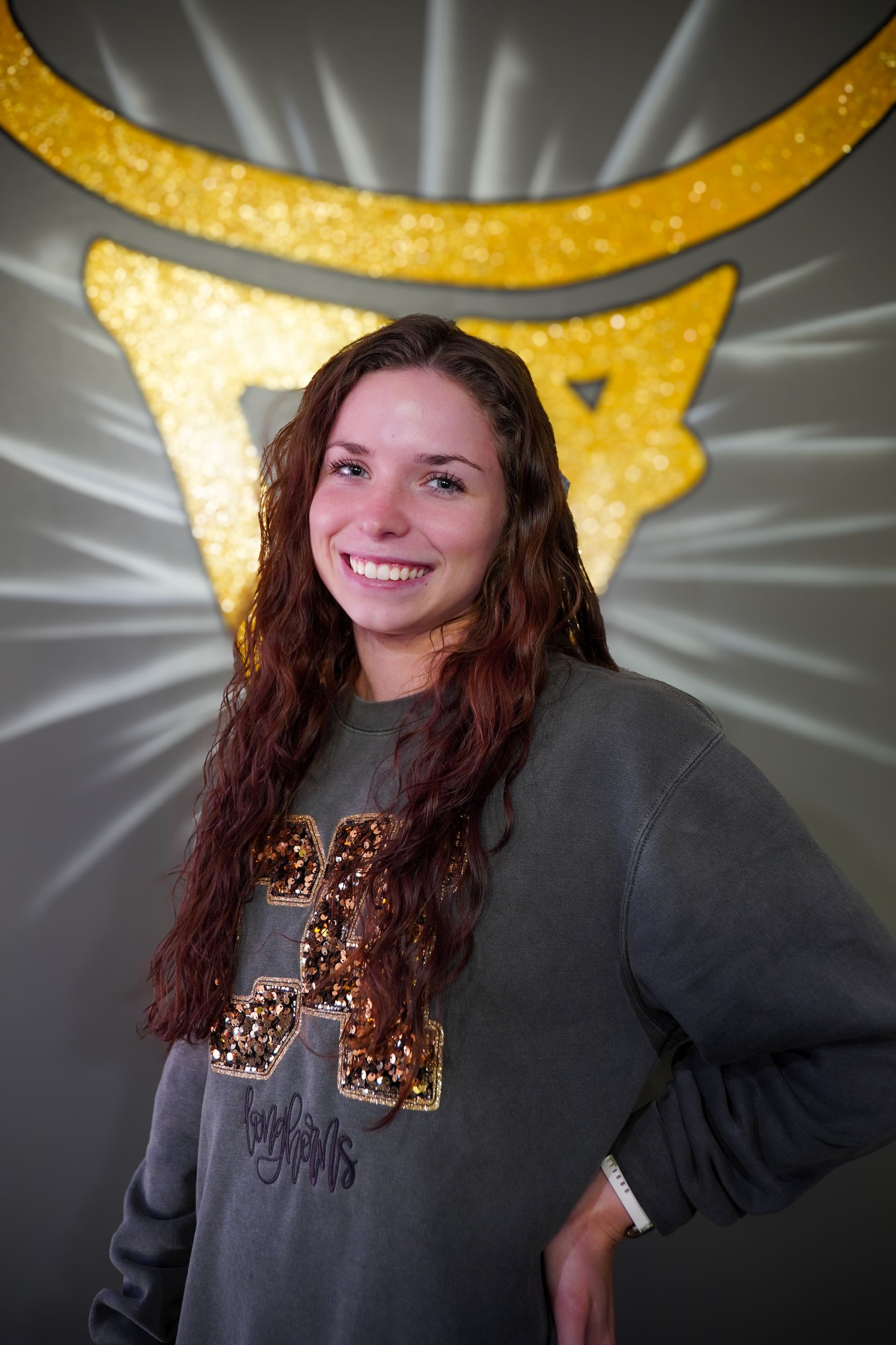 Woman with long curly hair smiles, wearing a green sweatshirt with an emblem in front of a golden logo on a gray wall.
