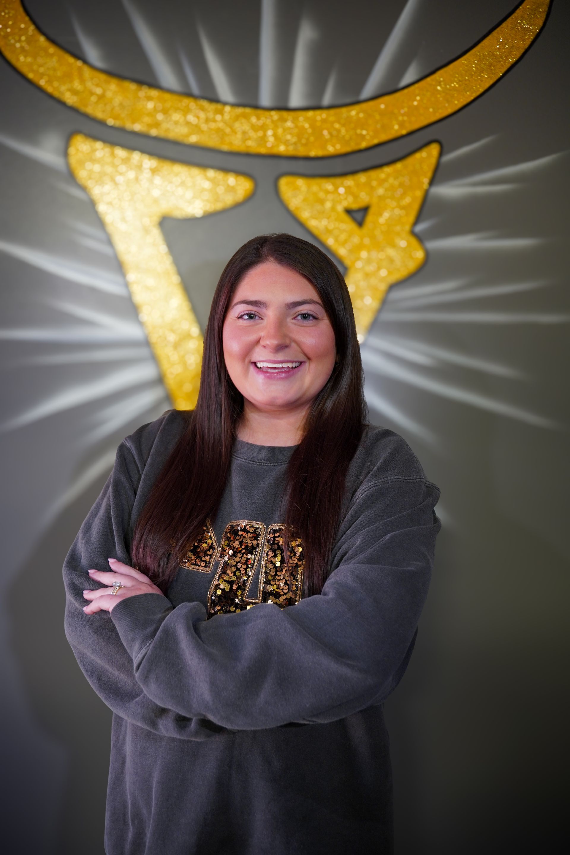 Woman with arms crossed, smiling, in front of a golden bull logo on a gray wall.