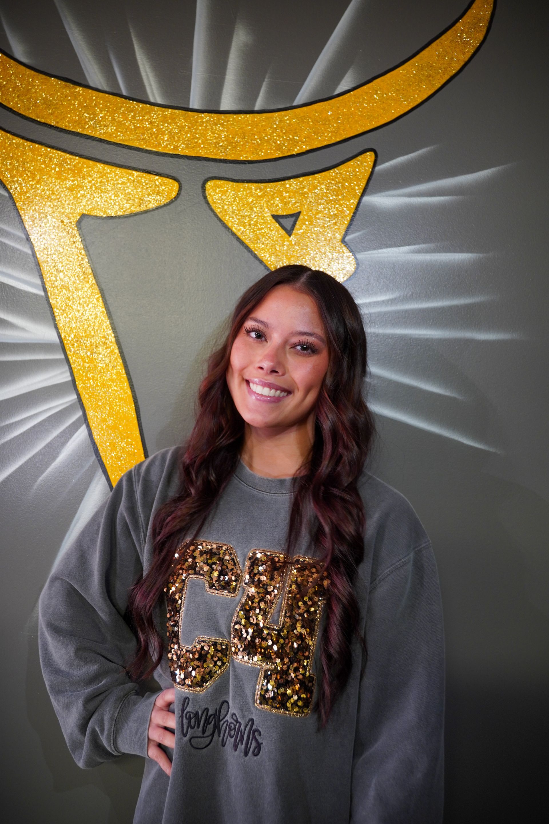 Woman smiling, wearing gray sweatshirt with gold lettering, standing in front of a wall with a gold design.