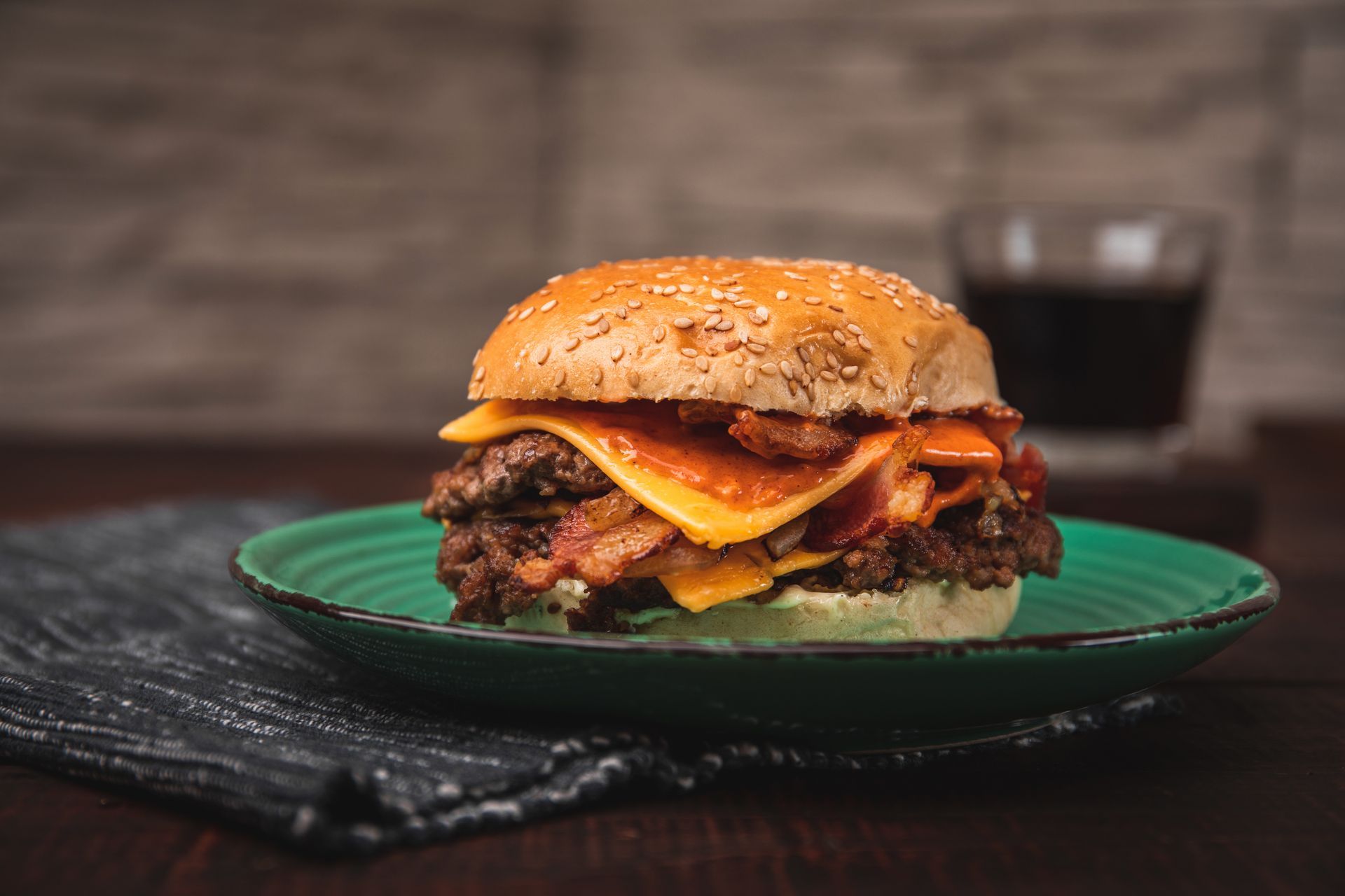 Double cheeseburger with bacon on a green plate, with a glass of dark liquid in the background.
