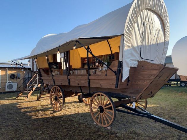 A wooden, covered wagon-style outdoor bar with string lights, set on a grassy field under a clear blue sky.