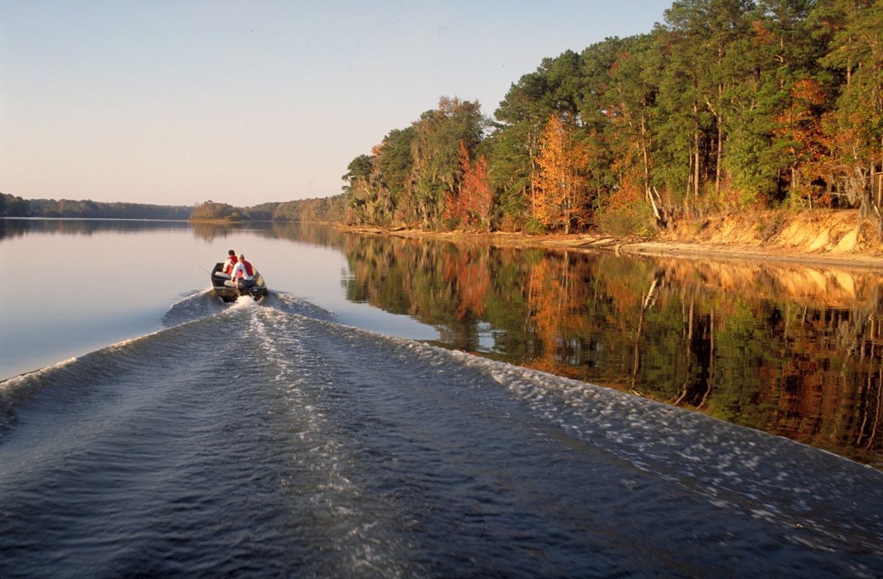 A boat carrying two people travels along a calm river lined with autumn trees, creating a wake in the water.