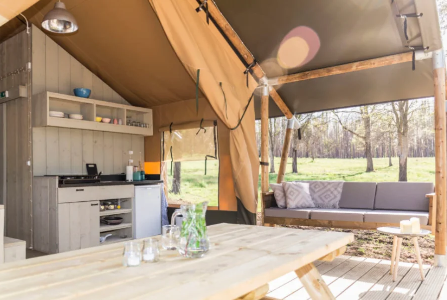An open-sided glamping tent featuring a kitchen area, a wooden dining table, and a sofa overlooking a wooded outdoor area.