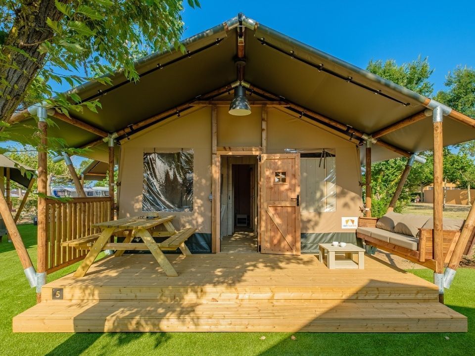 A large glamping tent with a wooden deck, outdoor dining table, and porch bench on a grassy site under a blue sky.