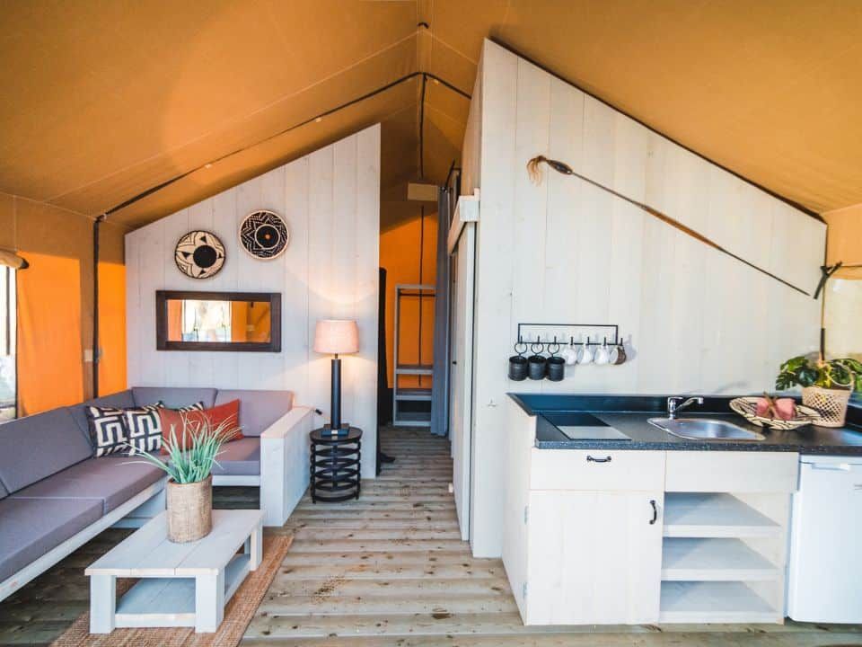 A modern, rustic-style tent interior with a gray sofa, coffee table, and kitchenette against white wooden walls.