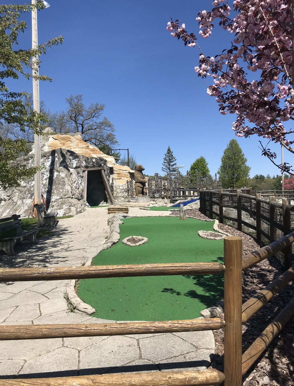 Miniature golf course on a sunny day with stone structures, green turf, wooden fences, and a blossoming pink tree.