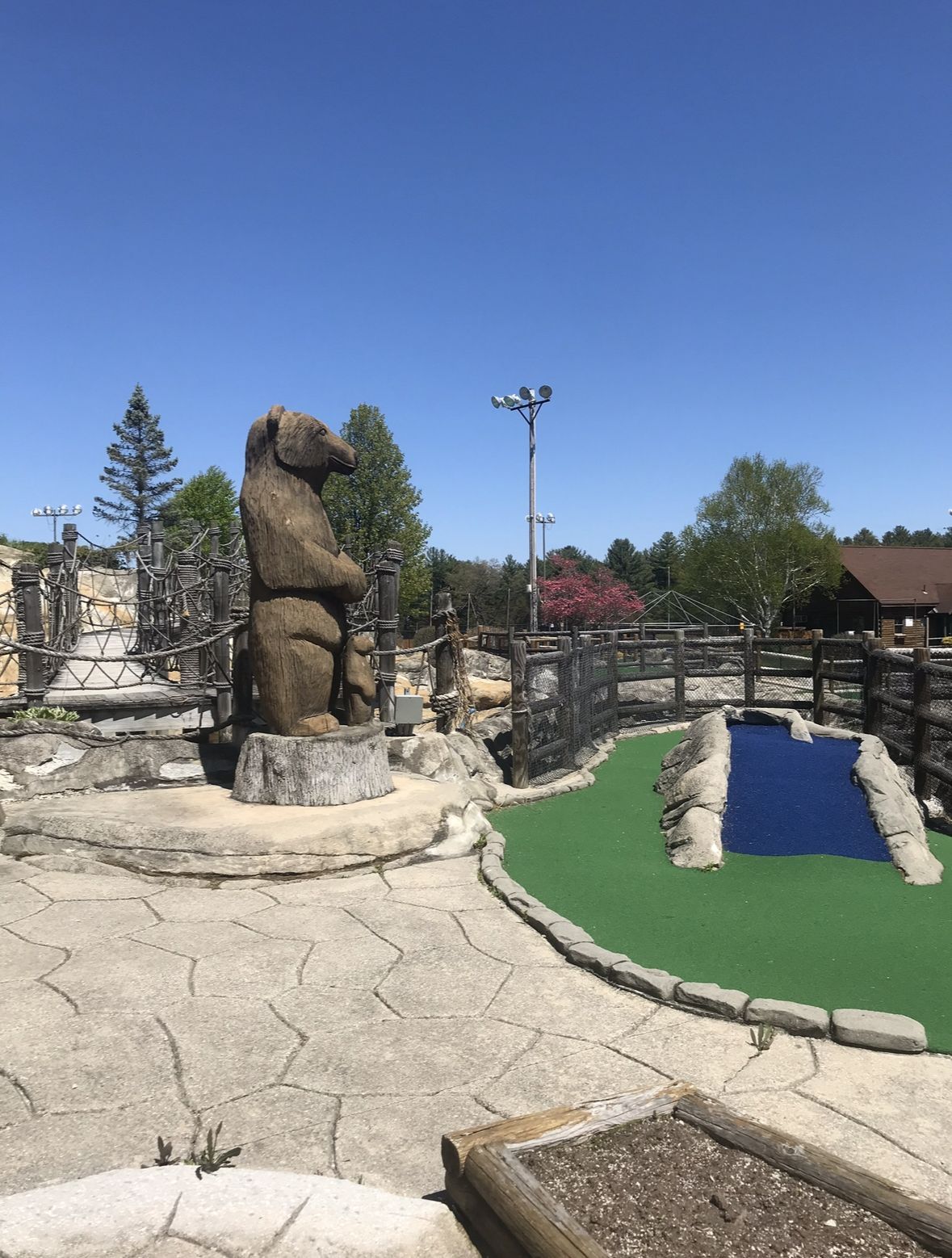 Mini golf course with a large wooden bear statue, green turf, and a blue water feature. Bright sunny day.
