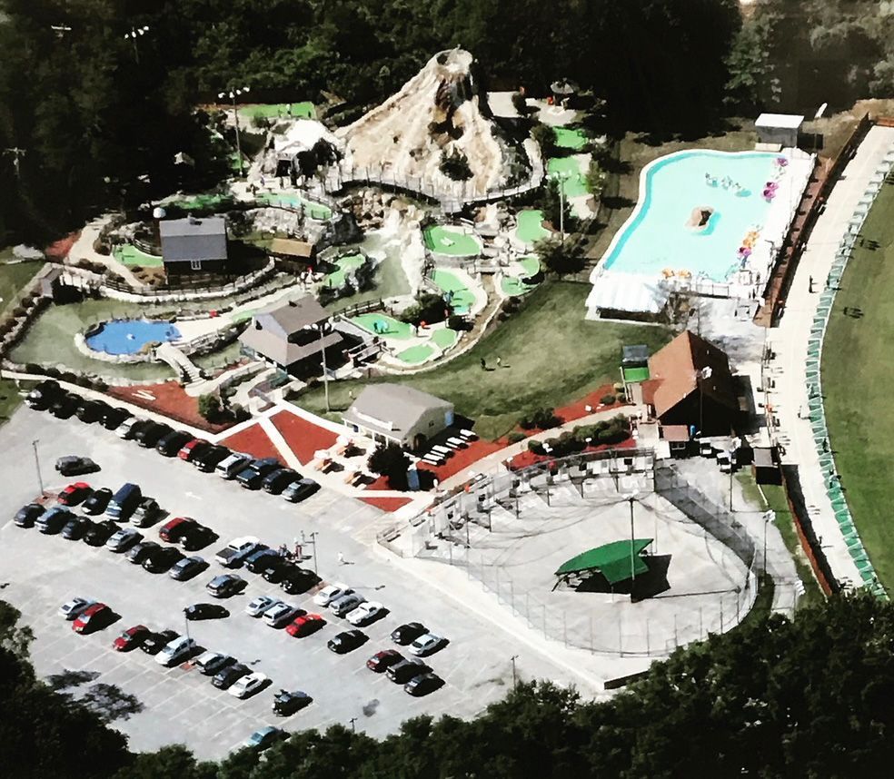 Aerial view of an amusement park with a volcano-themed miniature golf course, a pool, and a large parking lot.