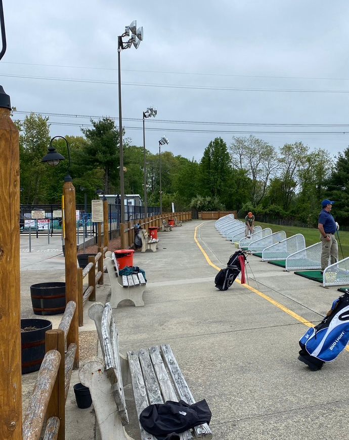 Golf driving range with benches, bags, and light poles. A person is walking down the lane.