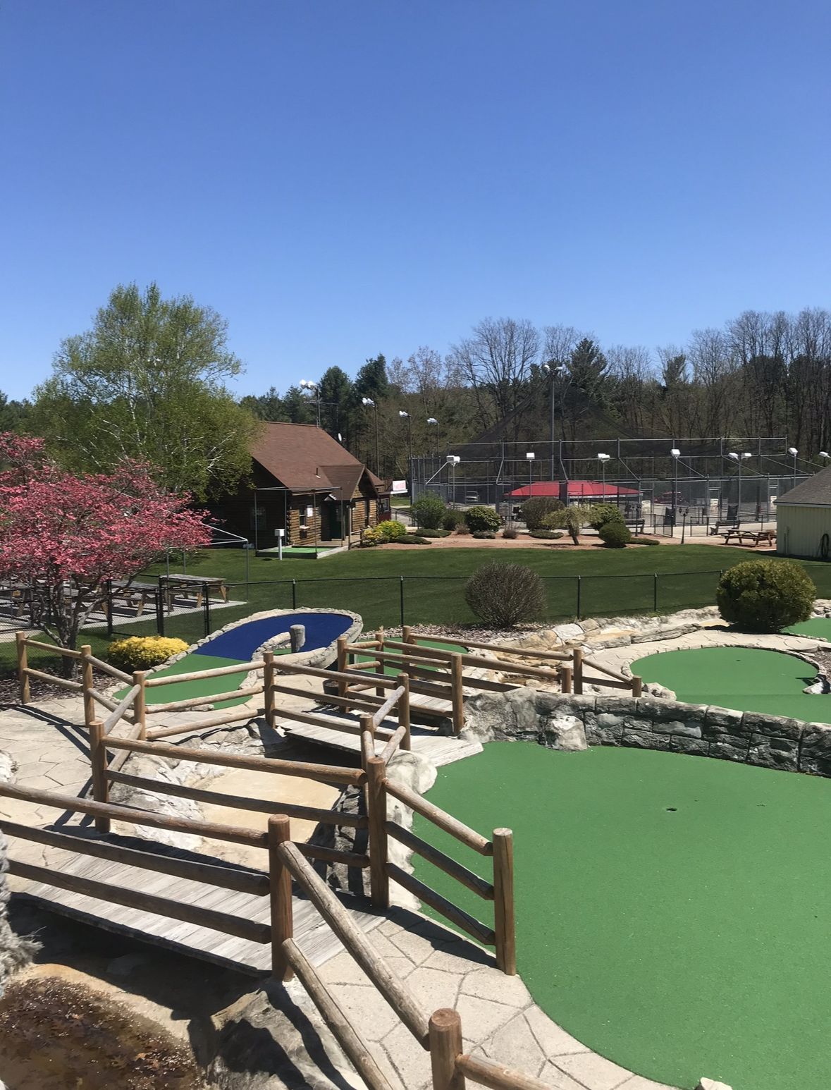 Miniature golf course with wooden bridges, green turf, blue water features, and a building on a sunny day.