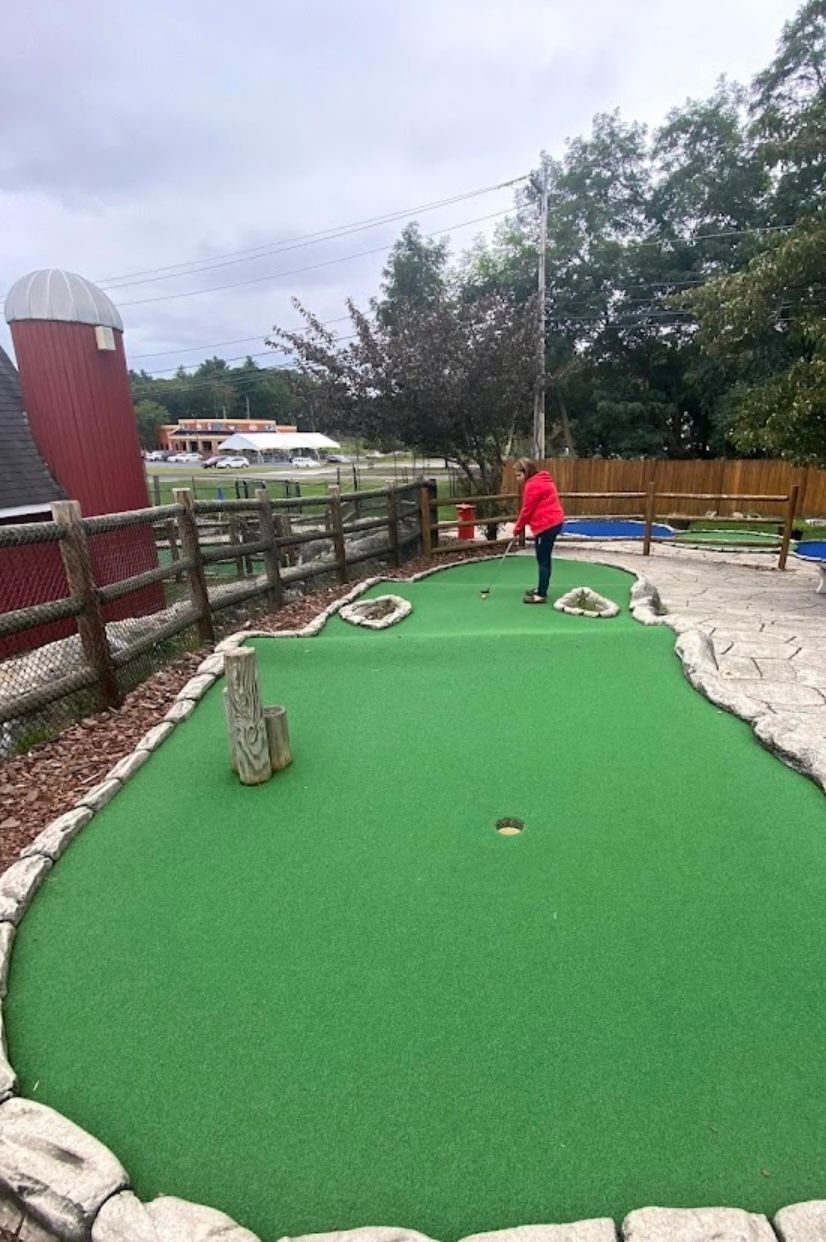 Person in red jacket playing mini golf on green course with a red barn in the background.