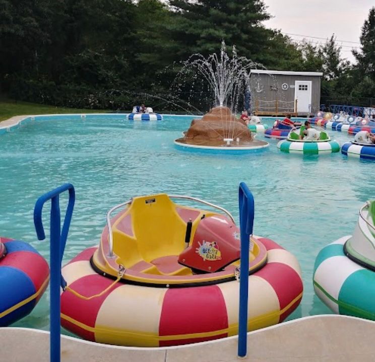 Bumper boats on a pool with a fountain and small building in the background. Boats are brightly colored.