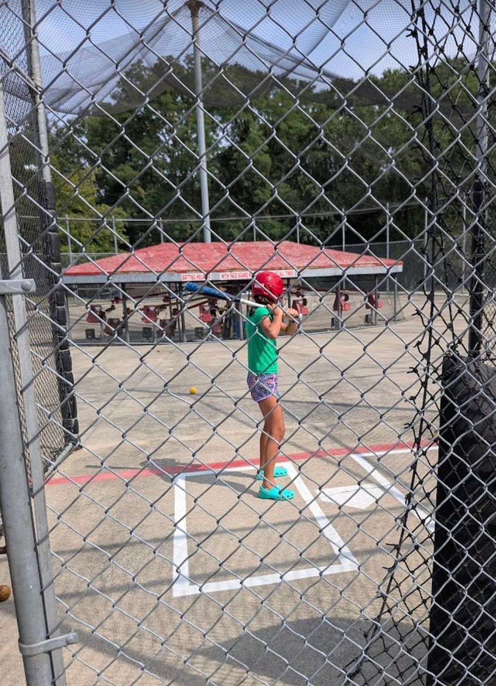 Girl in batting cage with bat, green shirt, red helmet, ready to swing. Cage in foreground, pavilion in background.