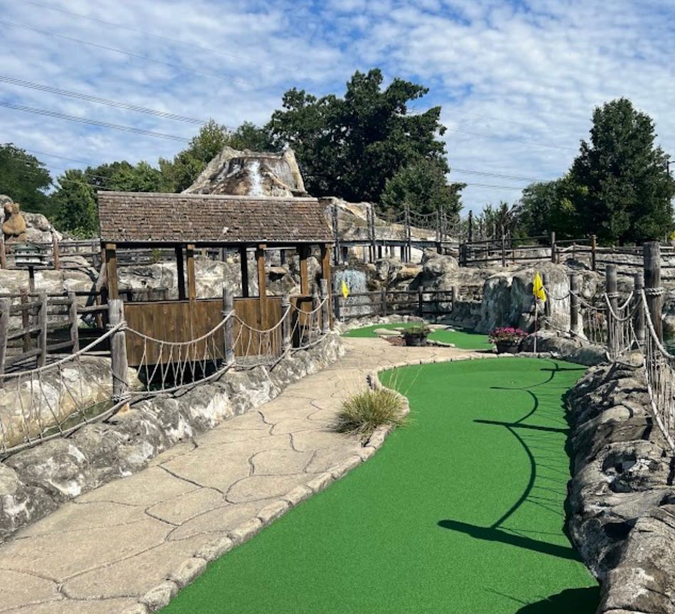 Miniature golf course with green turf, stone walls, and wooden structures under a cloudy sky.