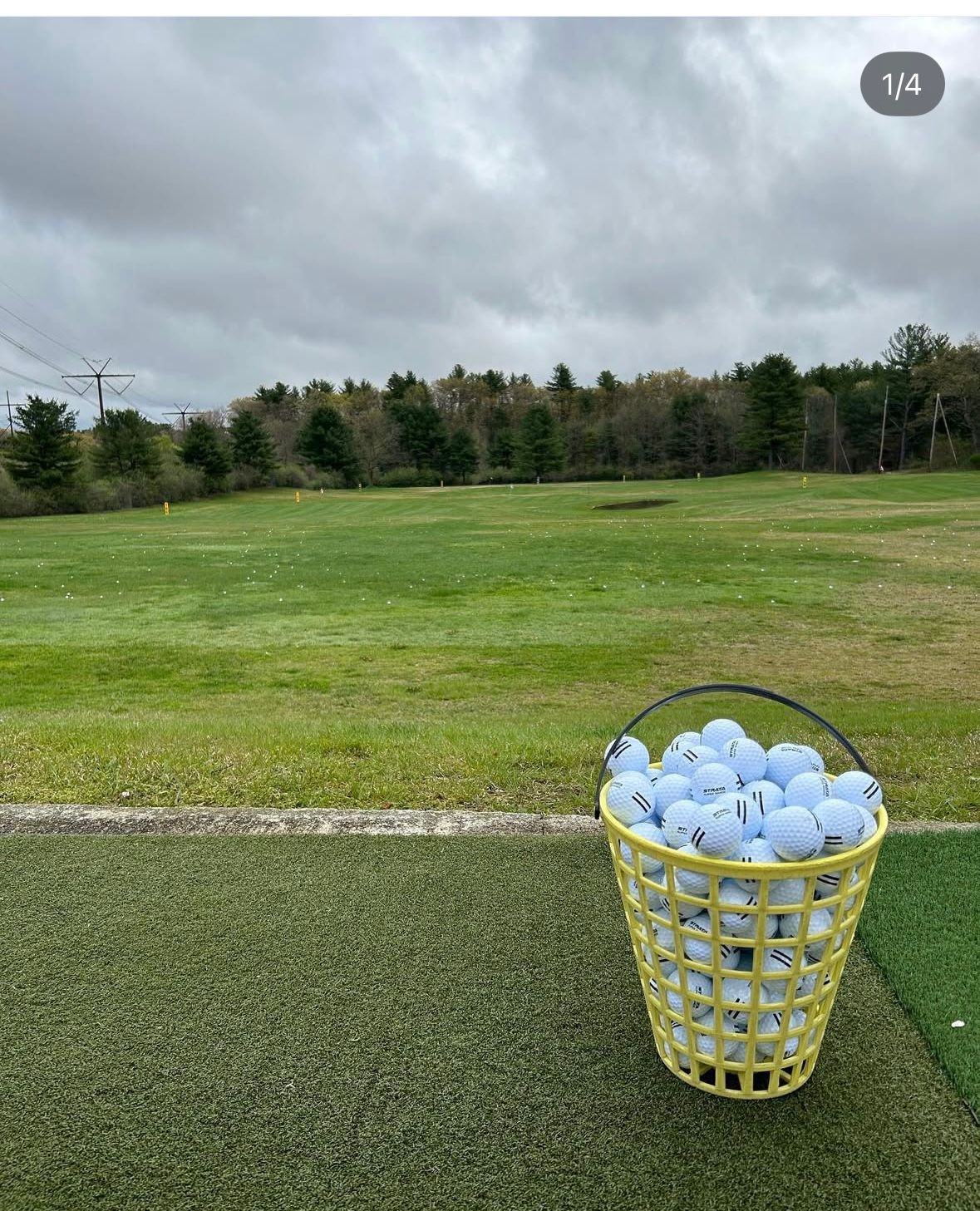 Bucket of golf balls on a driving range; green grass, trees, and cloudy sky in the background.