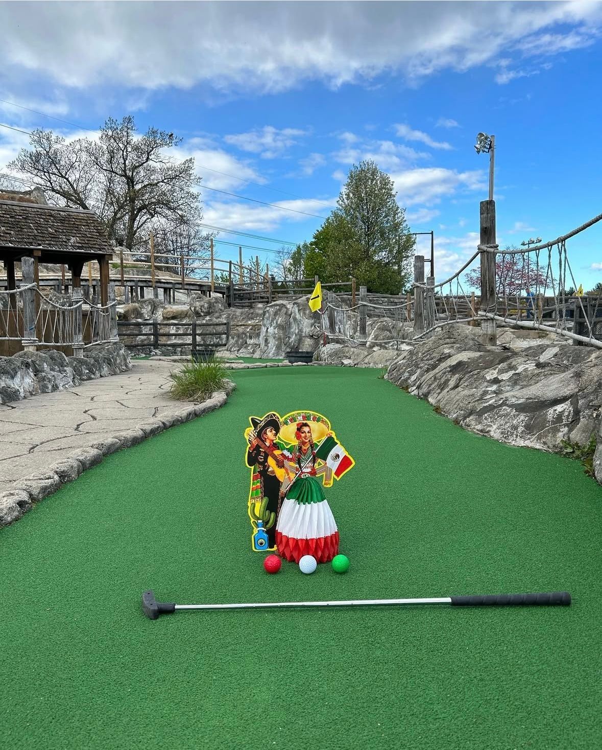 Miniature golf course hole with a festive decoration of a Mexican woman, a putter, and balls.