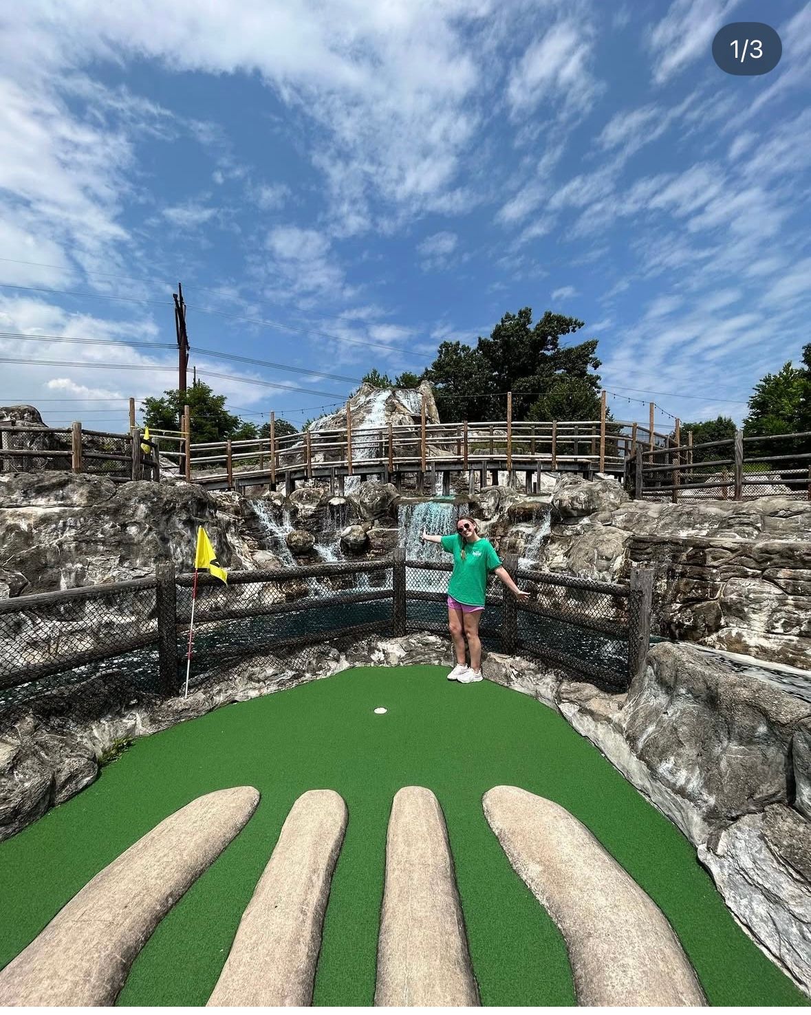 Woman standing on mini-golf green with a waterfall backdrop, blue sky overhead.