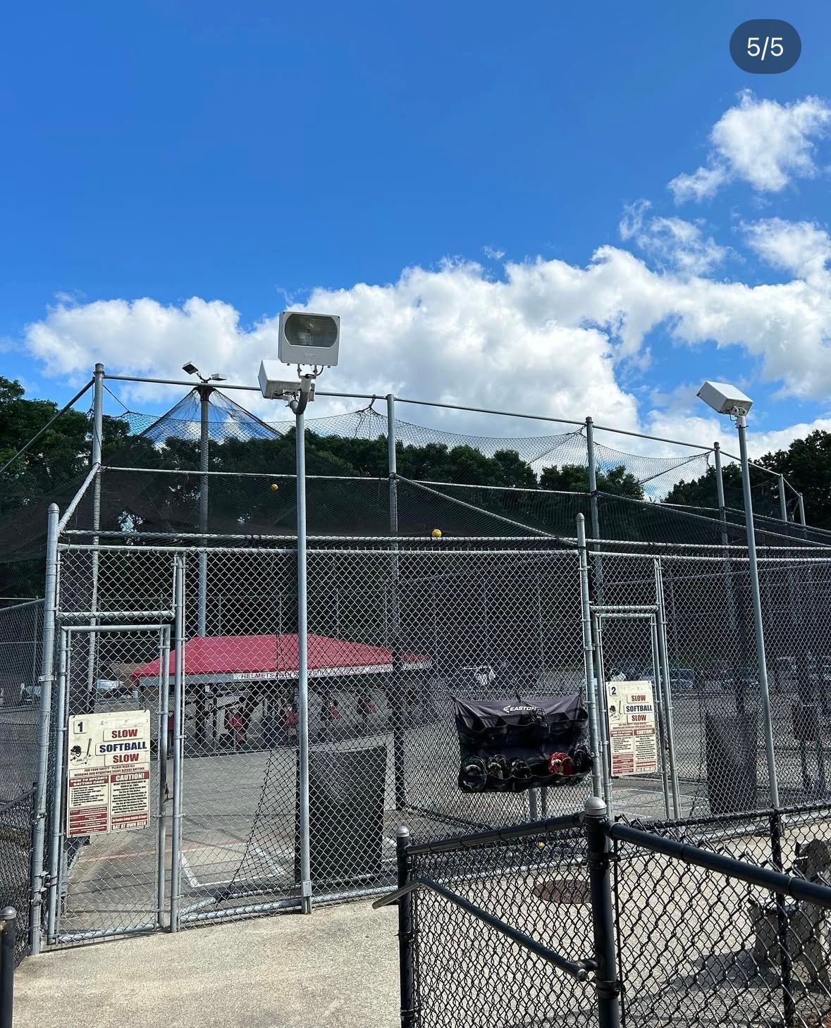 Fenced enclosure with overhead lighting, a clear blue sky, and a glimpse of red roof structure.