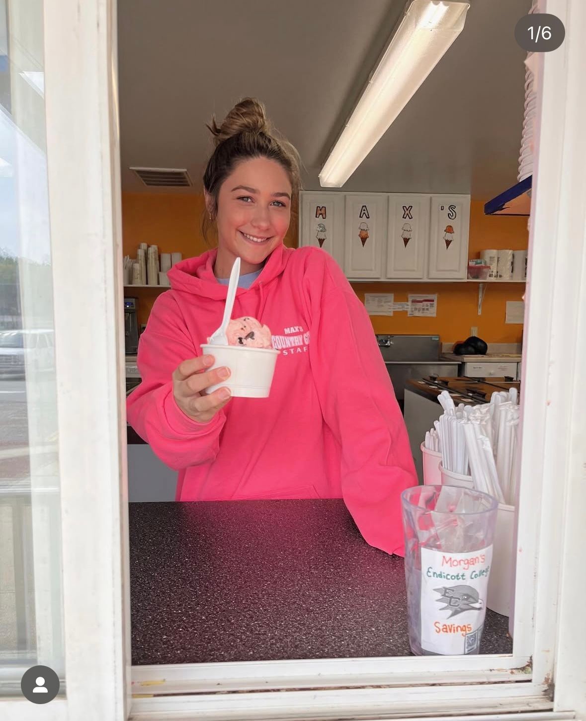 Woman in pink hoodie holding ice cream, smiling. Inside ice cream shop, serving customer.