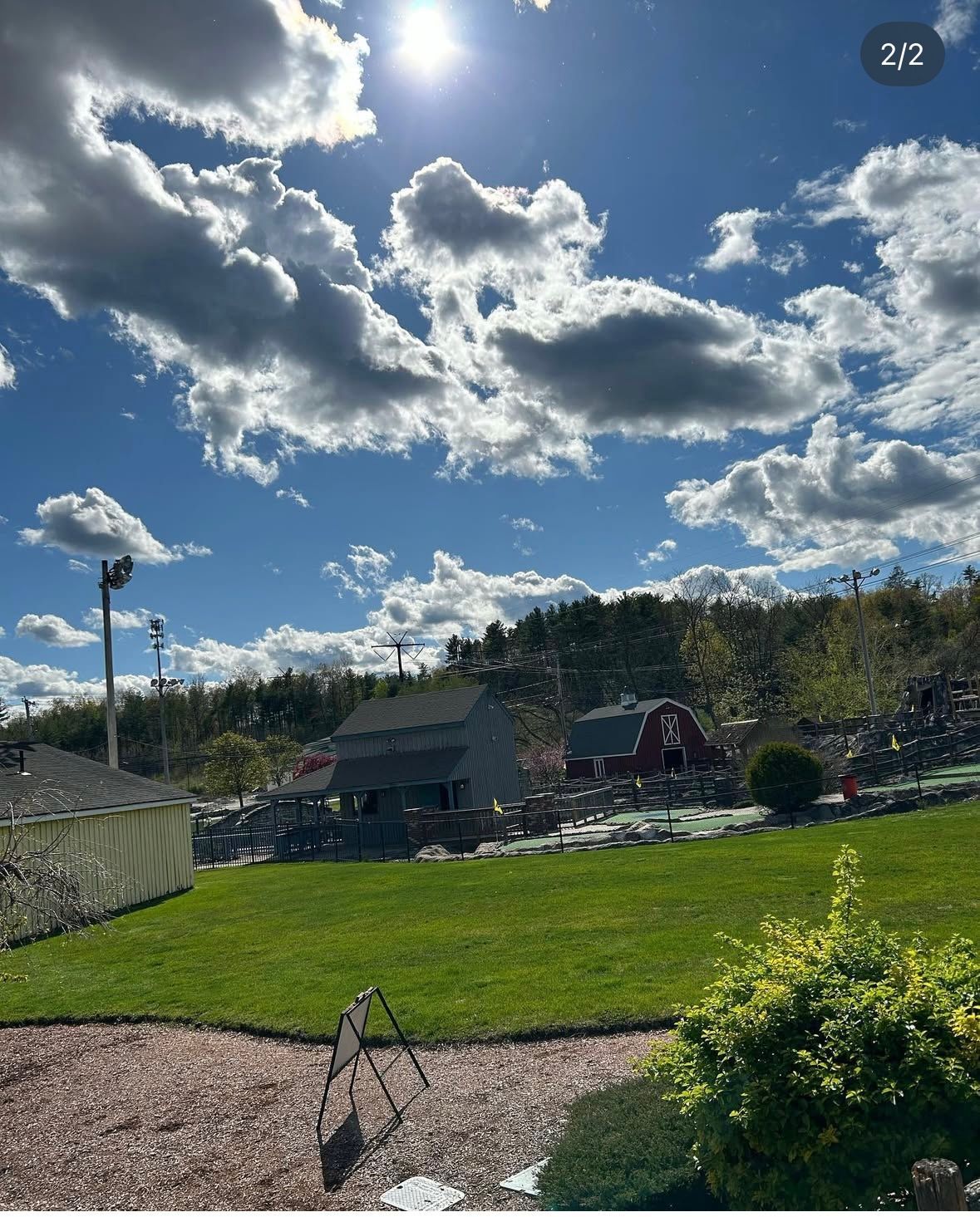 Sunny day with miniature golf course; blue sky, puffy clouds, green grass, and small buildings.