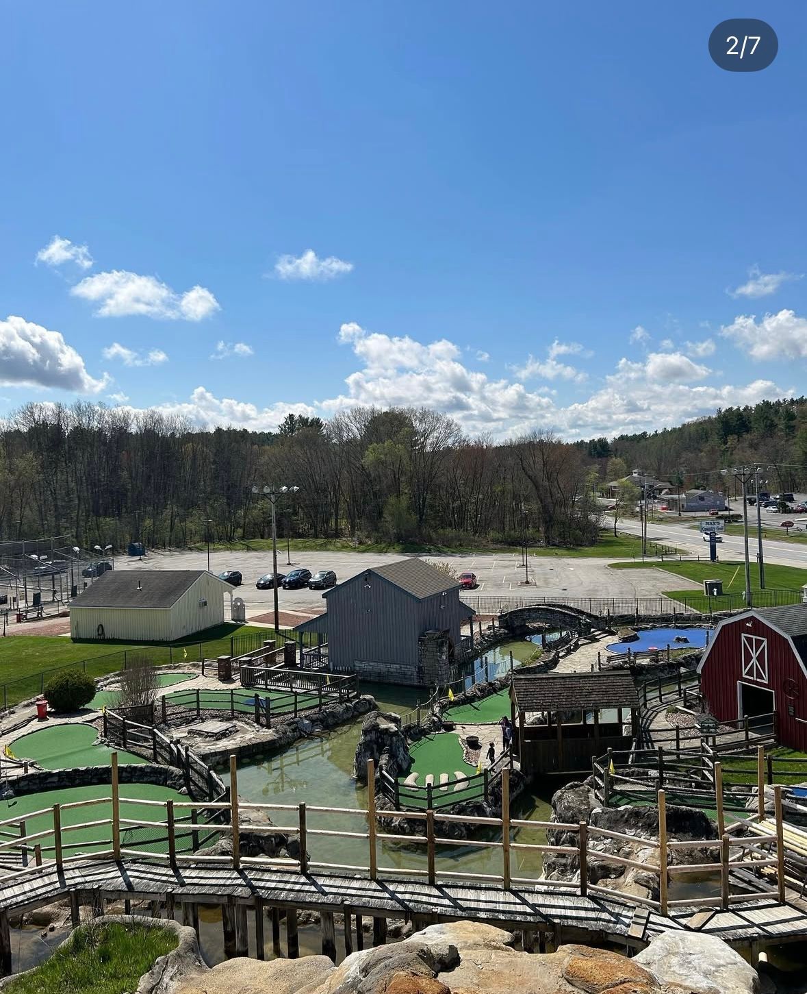Mini golf course with bridges, water features, and a red barn against a backdrop of trees, parking lot, and blue sky.