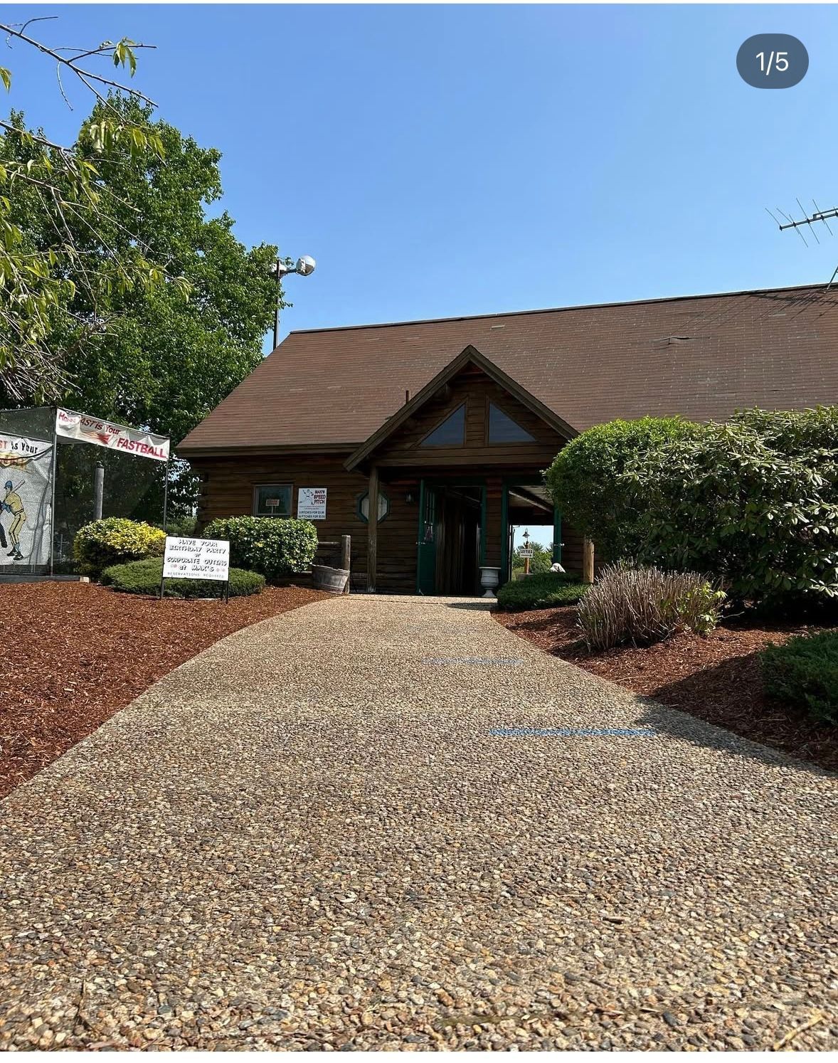 Log cabin building with gravel path, surrounded by shrubs, under blue sky.