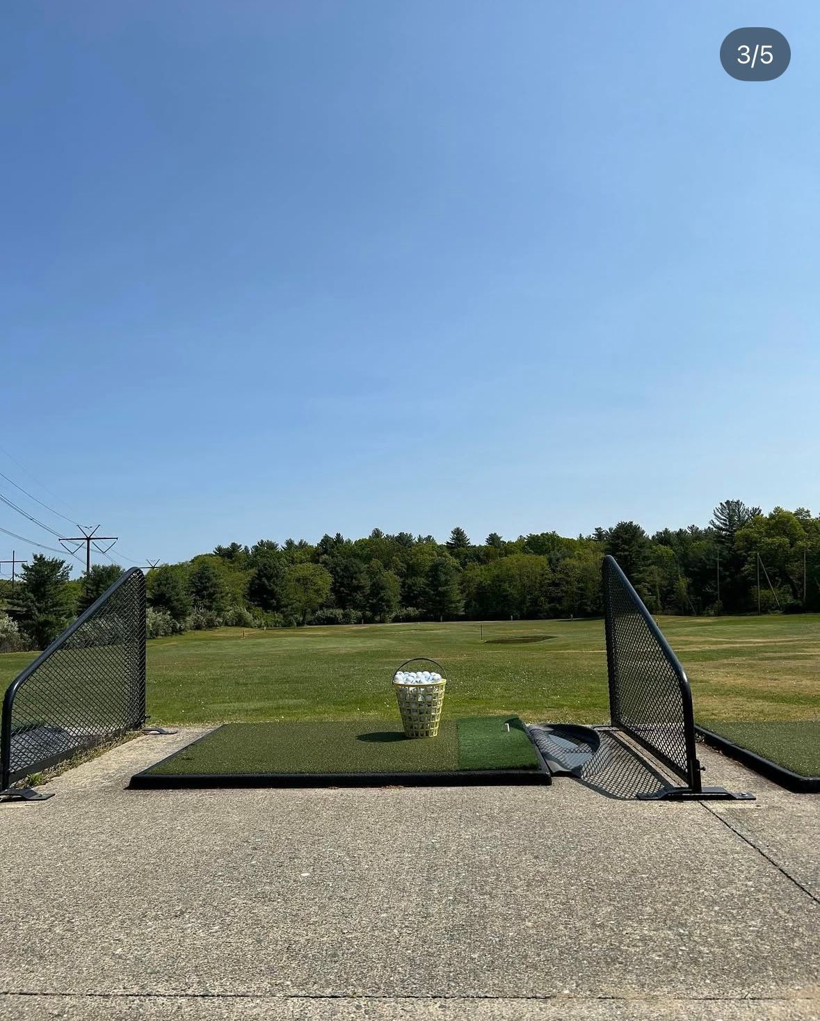 Golf driving range with a bucket of golf balls on a green mat, with a blue sky background.