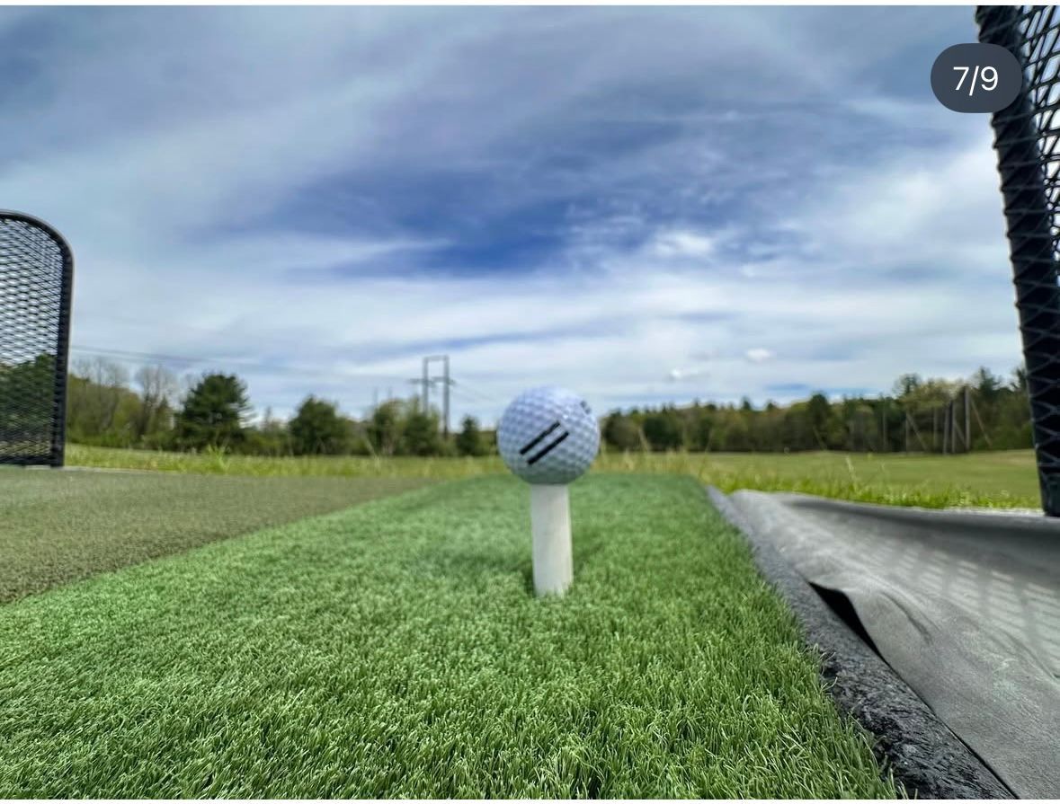 Golf ball on a tee, ready for a swing, set on a green artificial turf against a cloudy sky.