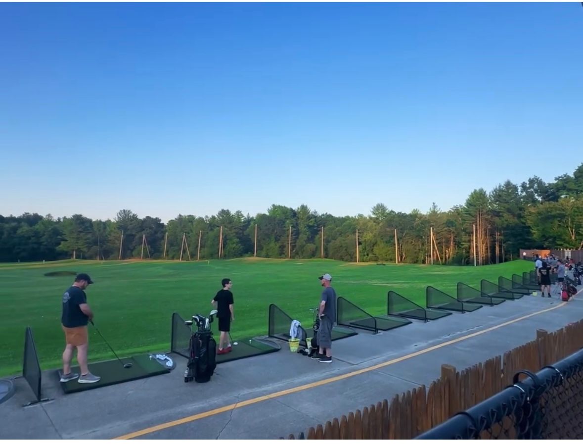 People practicing golf at a driving range, with green field, trees, and blue sky in the background.