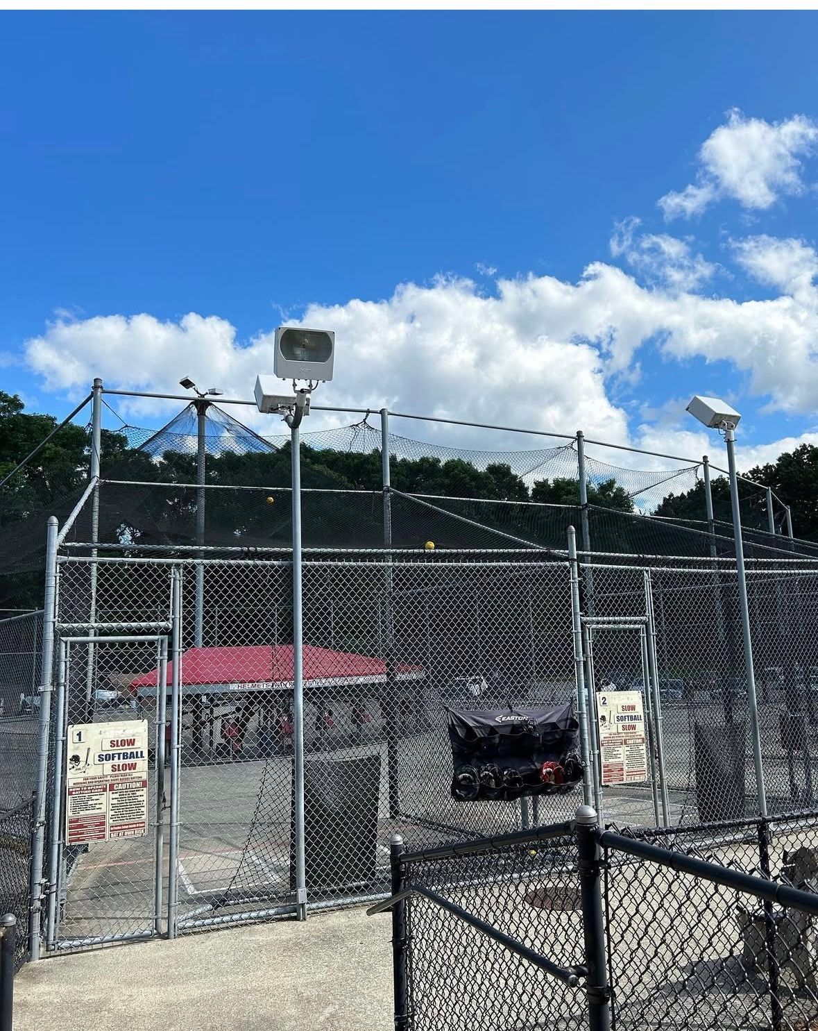 Fenced baseball batting cages with lights, under a cloudy blue sky, a building and trees visible in the background.