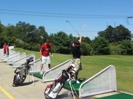 People practicing golf swings at a driving range. Green grass, blue sky, golf bags.