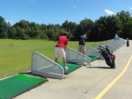 People practicing golf swings at a driving range, sunny day.