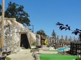 Miniature golf course with rock structures, a seated statue, and green turf against a blue sky.