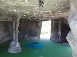 Mini-golf cave-themed hole with green turf, stone walls, and a blue ring. Daylight visible through the exit.