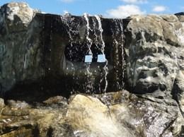 Waterfall cascading over artificial rocks with a square opening, against a blue sky.
