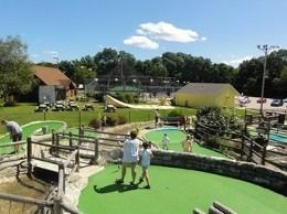 Mini golf course on a sunny day. People play, with green turf, wooden features, and a yellow building in the background.