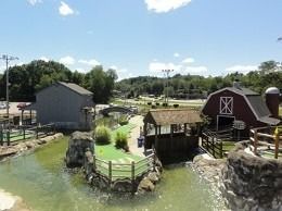 Miniature golf course with a red barn, water features, and green landscaping under a bright sky.