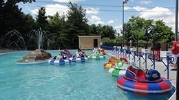 Bumper boats in a pool with a fountain, trees, and a light blue sky.