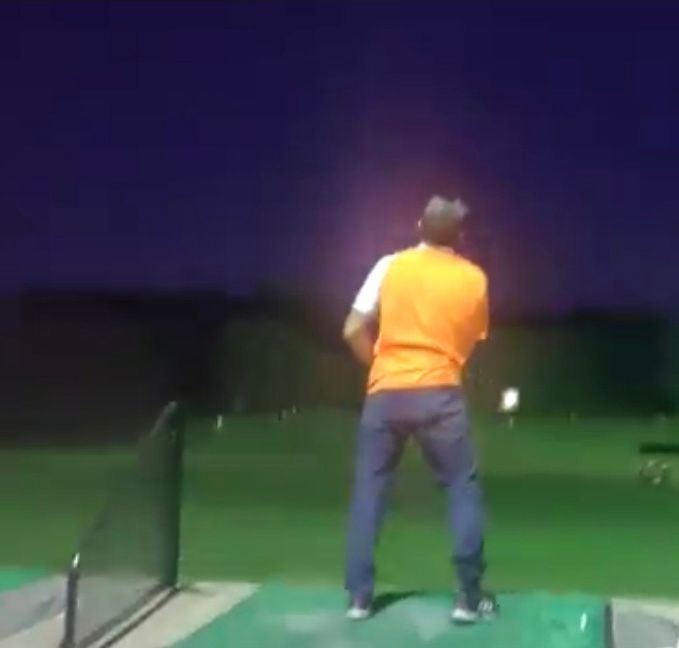 Man swinging a golf club at a driving range at night, wearing orange shirt and jeans.