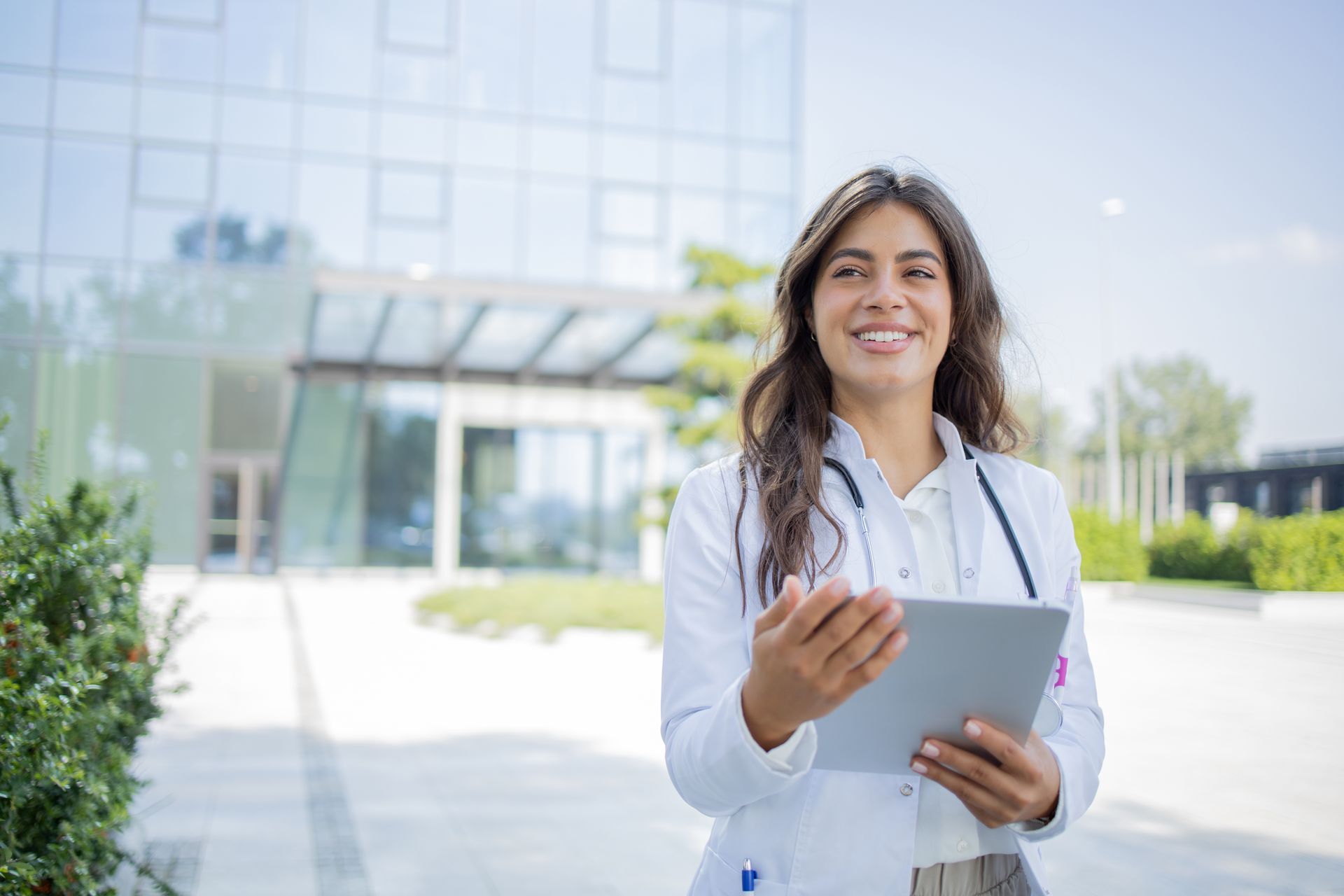 A female doctor is standing in front of a hospital holding a tablet.