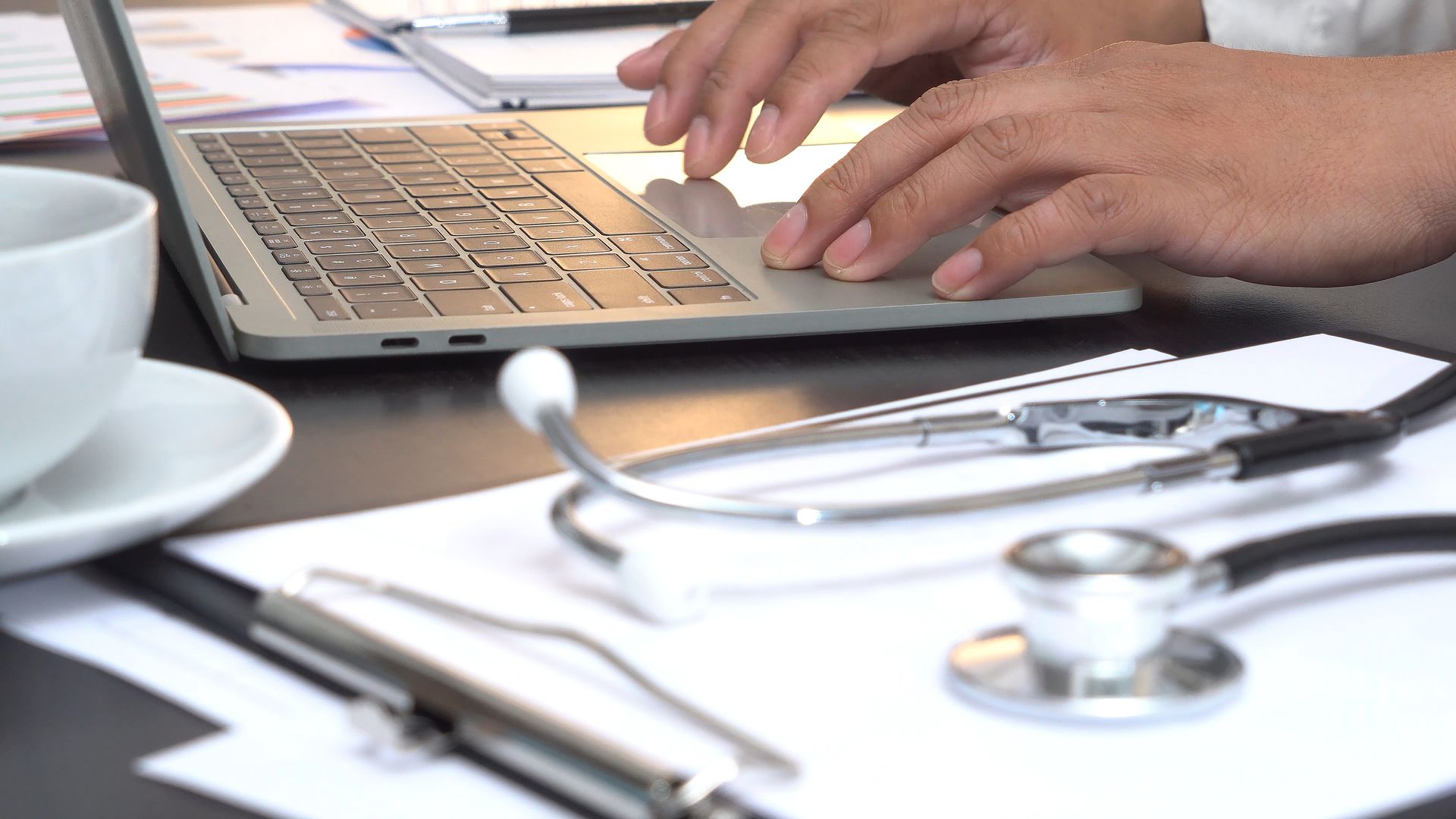 A doctor is typing on a laptop computer next to a stethoscope.