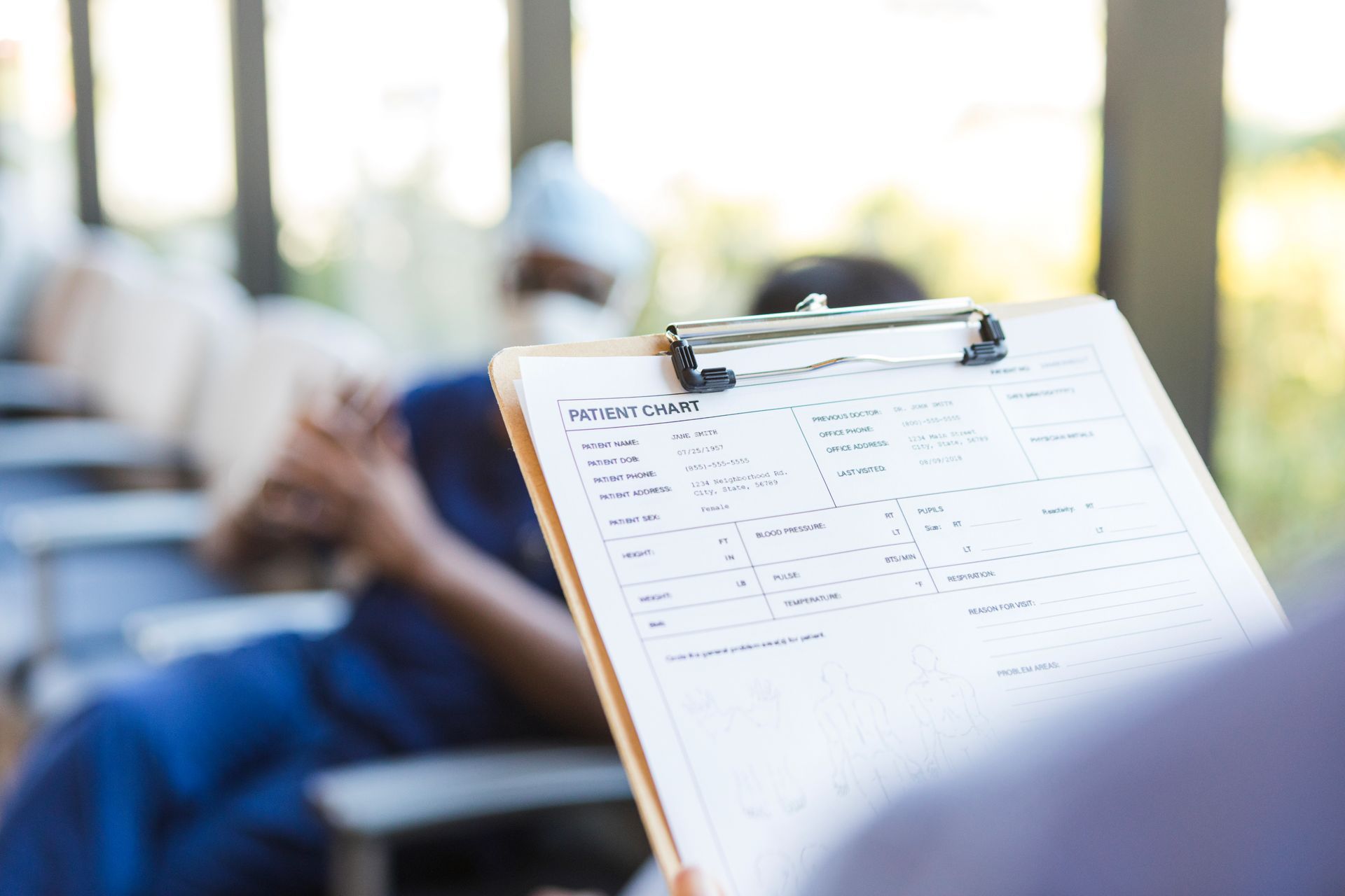A person is holding a clipboard with a patient sitting in the background.