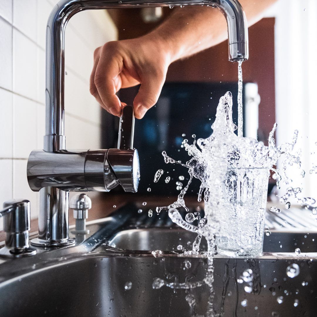 A hand turns on a faucet, water splashing into a glass in a stainless steel sink.