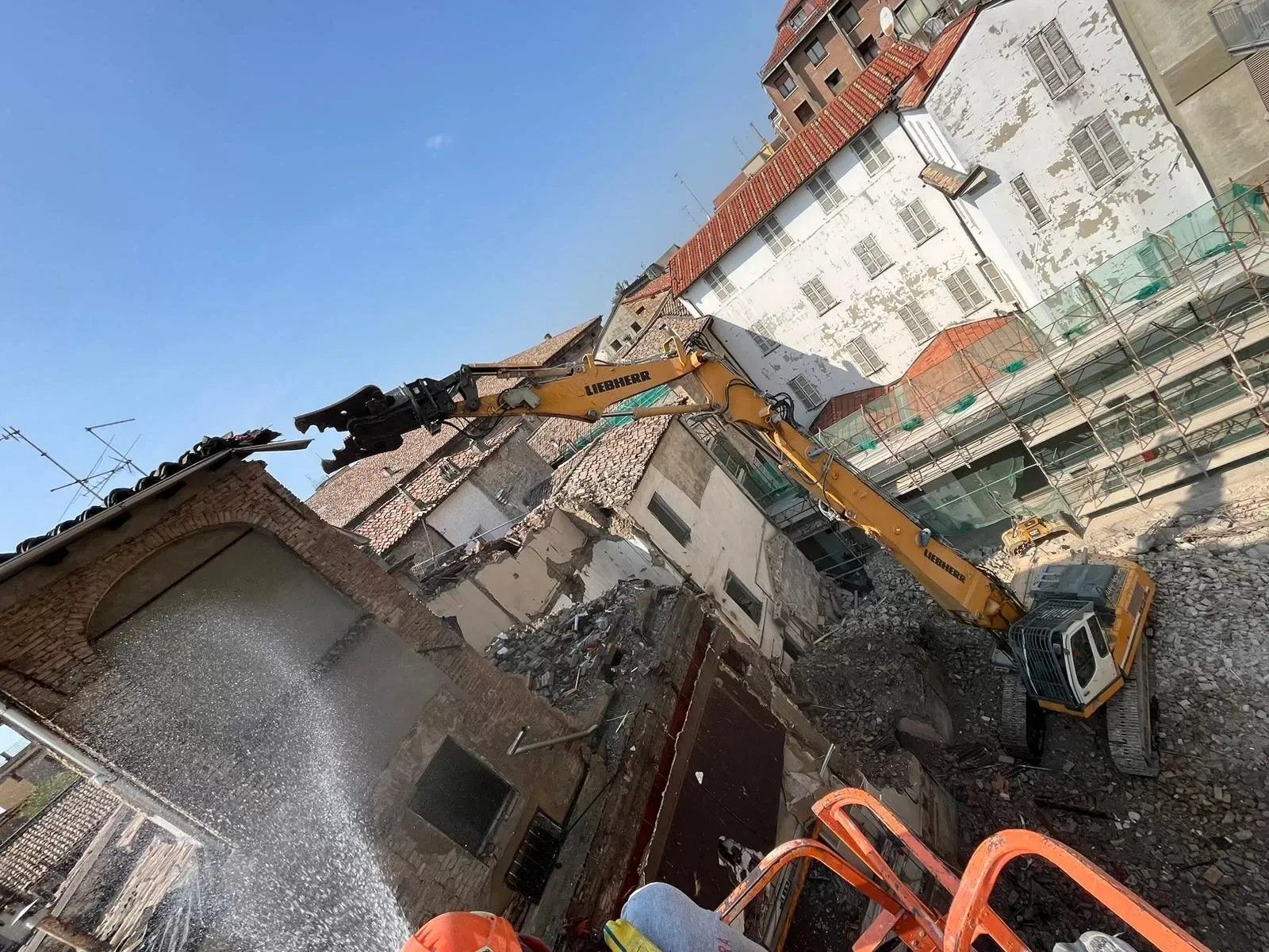 Un escavatore giallo demolisce un edificio in mattoni sotto un cielo azzurro e limpido.