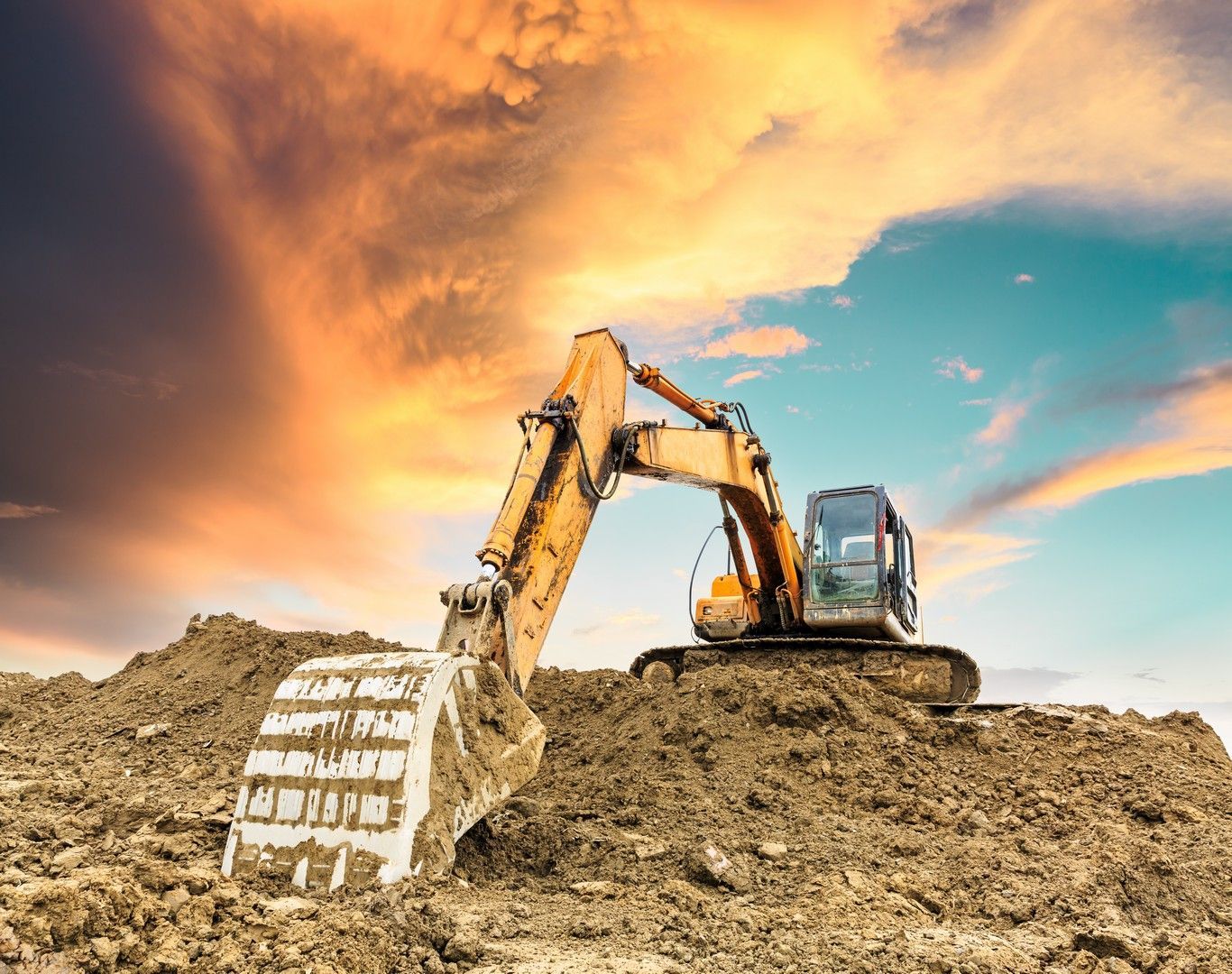Un escavatore giallo si erge su un cumulo di terra, stagliandosi contro un cielo luminoso e nuvoloso al tramonto.