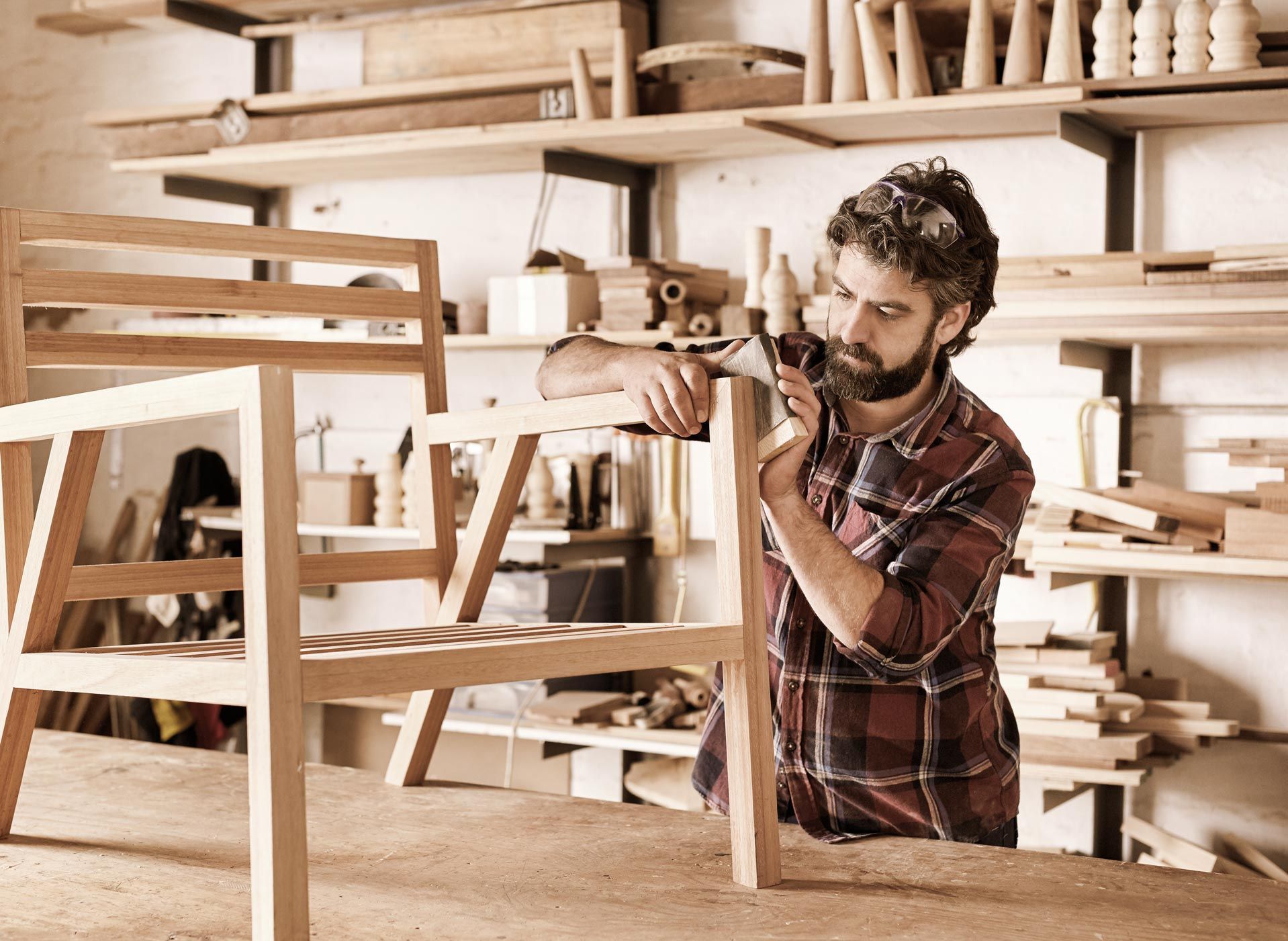 Carpenter inspecting wooden chair in workshop, tools and shelves in the background.