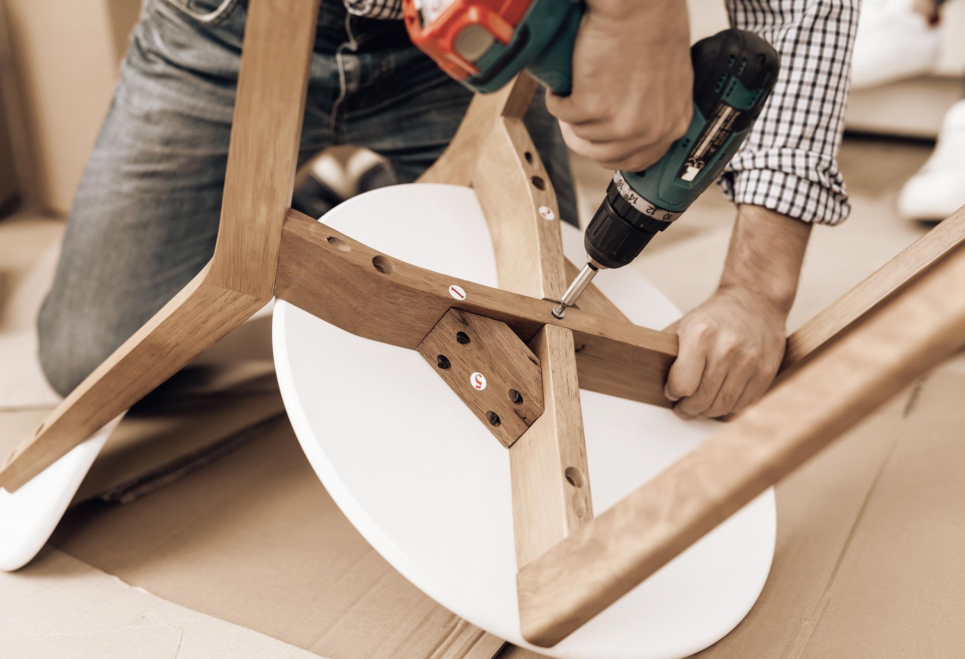 Person using a power drill to assemble a white and wooden chair on cardboard.