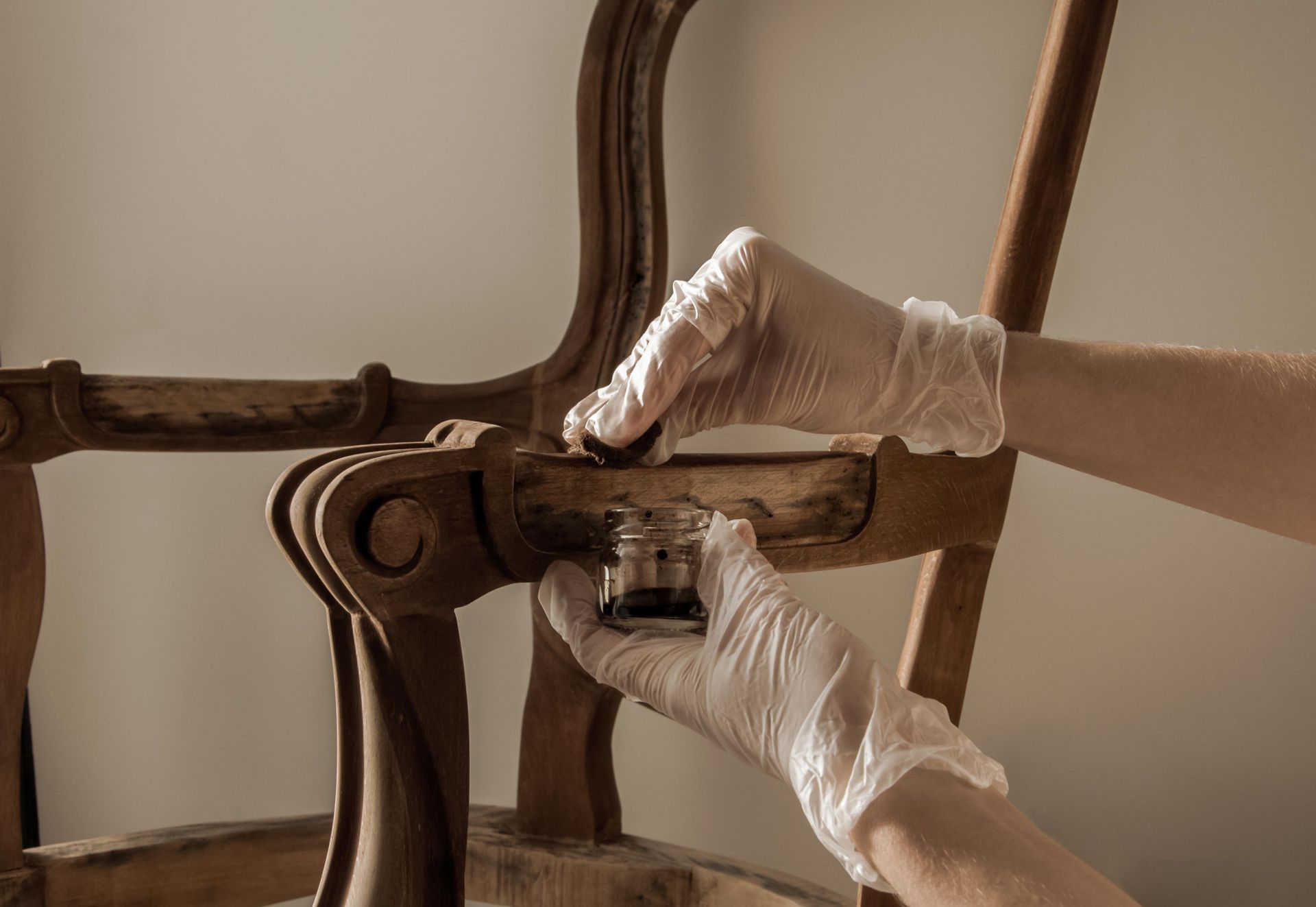 Hands in gloves applying stain to an antique wooden chair, indoors.